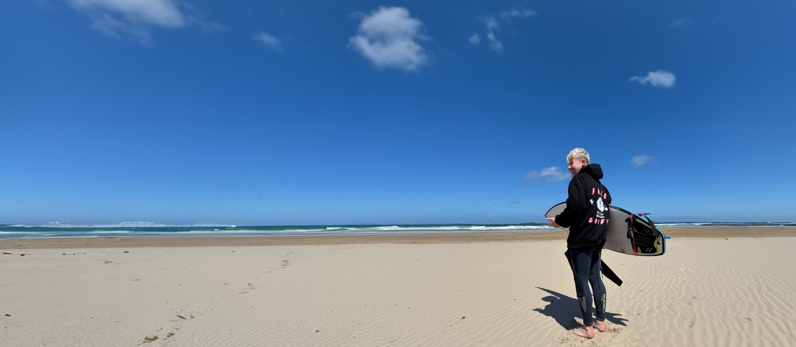 Surfer wearing Free Goofy hoody standing on a beach and holding his surfer board.